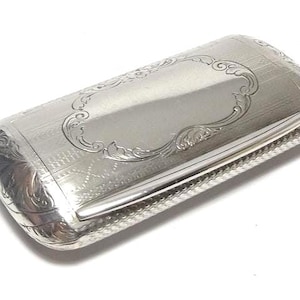 Puede incluir: Un estuche rectangular de metal plateado con bordes redondeados. La superficie superior presenta un cartucho central grabado rodeado de intrincados adornos y un patrón texturizado. Los lados y extremos están decorados con grabados florales detallados.
