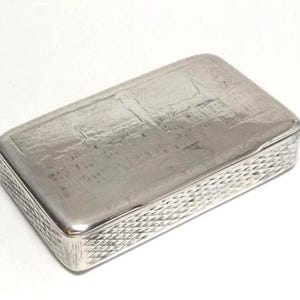 Puede incluir: Una caja rectangular de metal plateado con una escena de paisaje grabada en la tapa y un patrón de diamantes entrecruzados en los cuatro paneles laterales. La superficie tiene un acabado pulido y reflectante en todo el objeto.