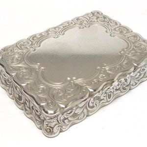 Puede incluir: Una caja rectangular de metal color plata con un panel central liso y pulido en la tapa. Los bordes de la tapa y los lados de la caja presentan intrincados grabados en relieve de volutas y motivos florales con un borde ondulado.