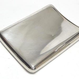 Puede incluir: Una pitillera rectangular de metal plateado con un acabado liso y pulido y esquinas redondeadas. El estuche cuenta con una tapa con bisagras y una sutil hendidura en el borde frontal para abrir la superficie brillante y reflectante.