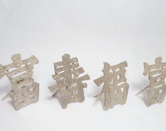 Op de afbeelding: Vier zilverkleurige Chinese karakterornamenten. Elk karakter is ingewikkeld ontworpen met een textuuroppervlak en staat rechtop met een kleine pin aan de basis. De karakters zijn in een rij gerangschikt tegen een witte achtergrond.