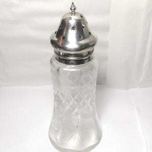 May include: A clear glass salt shaker with a silver-coloured metal lid. The glass has a diamond pattern and a flared base. The lid has small holes and a decorative finial. A classic kitchen or dining accessory.