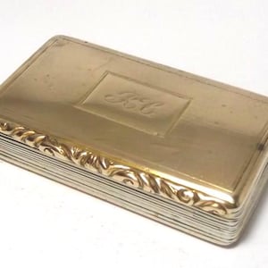 Puede incluir: Una caja rectangular de metal en tono dorado con tapa abatible. La superficie superior presenta un panel rectangular central grabado con las iniciales "KC" en letra cursiva. Los lados de la tapa están decorados con un patrón de volutas en relieve.