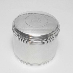 May include: A silver-coloured, round container with a lid. The lid has a slightly domed shape with a decorative emblem in the centre. The container has a hammered texture on the lid and a smooth, polished finish on the body.