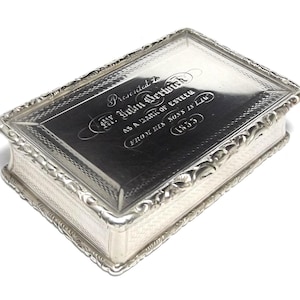 Op de afbeelding: Een rechthoekige zilveren doos met een scharnierend deksel. Het deksel is gegraveerd met tekst, waaronder de naam "Mr. John Berwick" en het jaar "1835". De doos heeft decoratieve randen en een patroon aan de zijkanten.