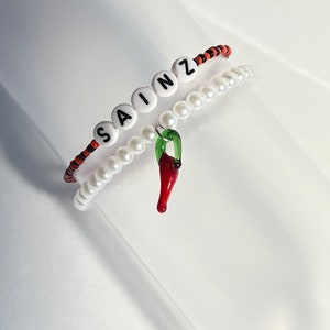 Może przedstawiać: Dwa bransoletki, jedna z białymi koralikami, a druga z czarnymi i pomarańczowymi koralikami. Bransoletka z białymi koralikami ma napisane imię "SAINZ" białymi literami na czarnych koralikach. Druga bransoletka ma zawieszkę w kształcie czerwonej papryczki chili.