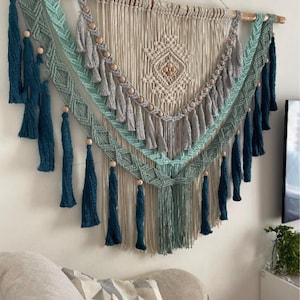 Könnte beinhalten: Ein Wandbehang aus Makramee mit einem geometrischen Muster in Beige, Hellgrün und Türkis. Der Behang ist mit Holzperlen und Quasten verziert.