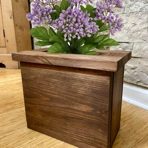 May include: A wooden planter box with a dark brown stain, holding a cluster of purple flowers with green leaves. The box has a rectangular shape and a simple, rustic design, suitable for indoor or outdoor use.