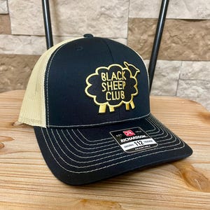 May include: Black and tan trucker hat with the text "BLACK SHEEP CLUB" in gold embroidery. The hat features a curved brim and a mesh back for breathability. The Richardson 112 hat is adjustable.