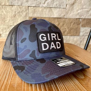 May include: A blue camouflage baseball cap with a black mesh back. The cap features a rectangular patch with the words "GIRL DAD" in white block letters. The brim has a Richardson 112 tag.