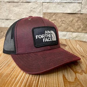 May include: A burgundy trucker hat with a black mesh back. The front features a black patch with the text "AIM FOR THE FACE" in white. The hat has a curved brim and a snap closure.
