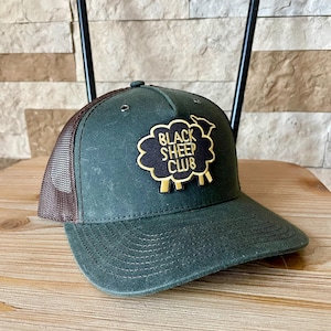 May include: A dark green baseball cap with a brown mesh back. The cap features a patch with the words "BLACK SHEEP CLUB" and a sheep illustration in black and gold.