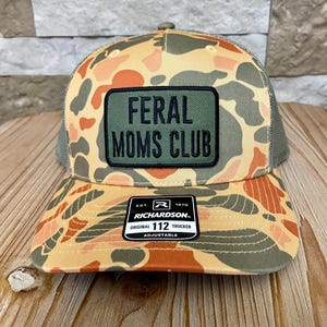May include: Camouflage trucker hat with a patch that reads "FERAL MOMS CLUB." The hat has a curved brim and a mesh back. The hat is adjustable and features the Richardson brand.
