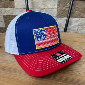 May include: A blue, white, and red trucker hat with a QR code design resembling a flag. The hat features a red brim, a white mesh back, and a blue front panel. The hat has the text "RICHARDSON" on a black tag.