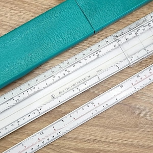 Puede incluir: Un conjunto de tres reglas de cálculo blancas con marcas negras y rojas, junto con un estuche verde turquesa. Las reglas de cálculo tienen escalas y medidas detalladas. El estuche es rectangular y de material rígido.