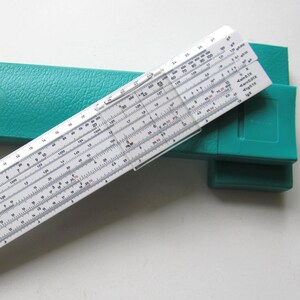 Puede incluir: Una regla de cálculo blanca con marcas negras y un cursor transparente, apoyada sobre una funda verde turquesa. La regla de cálculo es un instrumento científico antiguo utilizado para cálculos. La funda es rectangular y texturizada.