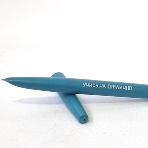 Puede incluir: Un bol&iacute;grafo de color turquesa con el texto "УЧИСЬ НА ОТЛИЧНО" impreso en el cuerpo. El bol&iacute;grafo est&aacute; sobre una superficie blanca, con la tapa separada. El bol&iacute;grafo tiene un dise&ntilde;o elegante.