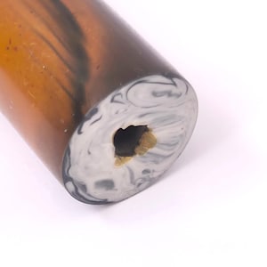 Puede incluir: Un objeto cilíndrico con un cuerpo liso y translúcido de color ámbar marrón con vetas oscuras y arremolinadas. La cara circular muestra un patrón jaspeado en tonos blancos, grises y negros con una abertura central irregular y oscura.