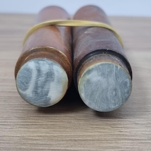 Puede incluir: Dos objetos cilíndricos de madera con un acabado liso y pulido, unidos por una banda elástica amarilla. Cada cilindro presenta una tapa circular con un patrón jaspeado de remolinos grises y blancos, mostrando una textura definida.