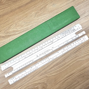 Puede incluir: Un conjunto de reglas de cálculo blancas con marcas negras, y una funda verde. Las reglas de cálculo están dispuestas sobre una superficie de madera. La funda es rectangular y tiene una superficie texturizada. Las reglas de cálculo tienen varias escalas y marcas.