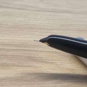 Puede incluir: Primer plano de una pluma estilogr&aacute;fica negra con plum&iacute;n plateado. La pluma est&aacute; inclinada hacia arriba a la izquierda, con el plum&iacute;n bien enfocado. El cuerpo de la pluma es liso y brillante, contrastando con la superficie de madera mate.