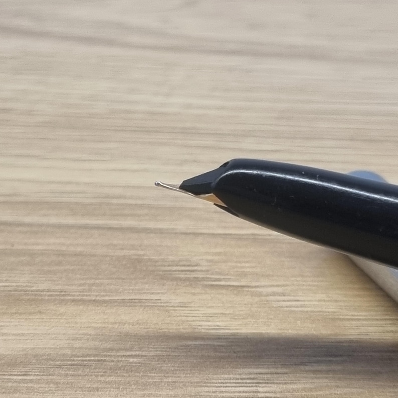 Puede incluir: Primer plano de una pluma estilogr&aacute;fica negra con plum&iacute;n plateado. La pluma est&aacute; inclinada hacia arriba a la izquierda, con el plum&iacute;n bien enfocado. El cuerpo de la pluma es liso y brillante, contrastando con la superficie de madera mate.