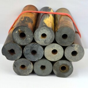Op de afbeelding: Een bundel van tien cilindrische staven gemaakt van gemarmerd harsmateriaal in tinten bruin, zwart en grijs. Elke staaf heeft een hol gat in het midden dat door de hele lengte loopt. De hele collectie is samengebonden met een dunne rode elastiek.