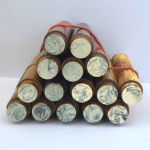 Puede incluir: Una colección de varillas cilíndricas de madera agrupadas con bandas elásticas rojas y verdes. Cada varilla presenta una tapa circular con un patrón jaspeado en tonos crema, gris y negro, que contrasta con los cuerpos de madera marrón lisa.