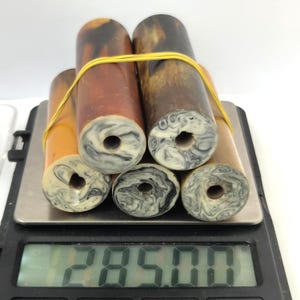 May include: Five cylindrical rods with amber-coloured bodies and marbled white and black ends, secured together by a yellow rubber band. The rods are resting on a digital scale that displays a weight of 285.00 grams, showing the total mass of the items.