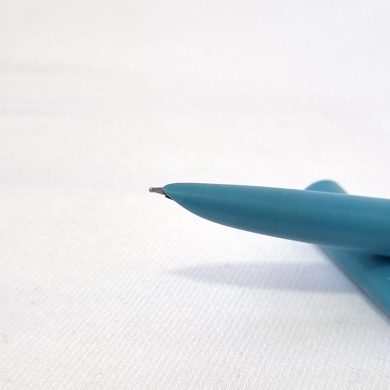 Puede incluir: Primer plano de una pluma estilogr&aacute;fica turquesa con plum&iacute;n plateado. La pluma es de color liso y tiene un dise&ntilde;o suave y redondeado. La pluma est&aacute; sobre una superficie blanca.