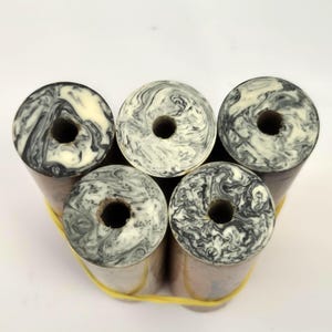 May include: Five cylindrical pen blanks arranged in a cluster and secured with a yellow rubber band. Each blank features a marbled pattern of black and white swirls on the top surface and a hollow circular centre hole running through the middle.