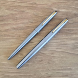 Może przedstawiać: Dwa szare długopisy ze złotymi akcentami są prezentowane na drewnianej powierzchni. Długopisy mają eleganckie, metaliczne wykończenie i klasyczny design. Długopisy są skierowane ku sobie, prezentując swoją elegancką formę.