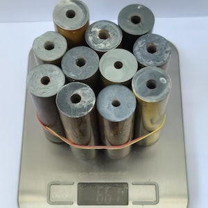 May include: A collection of eleven cylindrical resin blanks with marbled patterns in shades of grey, brown, and cream, each featuring a central hole. The group is secured by a thin orange rubber band and rests on a digital scale displaying 661 grams.
