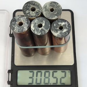 May include: Five cylindrical objects with copper-toned sides and marbled black and white circular tops, each featuring a central hole. The items are bundled together with a light-coloured rubber band and placed on a digital scale displaying 308.52 grams.