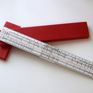 Puede incluir: Una regla de cálculo vintage con cuerpo blanco y marcas negras y rojas, sobre una funda roja. La regla de cálculo es una herramienta matemática para cálculos. La funda es rectangular y tiene una superficie texturizada.