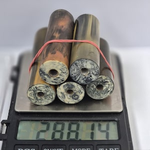 May include: Five cylindrical rods with a smooth, dark brown and tan finish, each featuring a circular hole in the centre with a marbled white and black pattern. The rods are bundled together with a thin red rubber band on a digital scale reading 288.14.