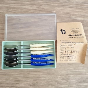 May include: A set of six small spoons in a light green plastic tray, with a clear plastic lid. The spoons have silver-colored bowls and handles with cream and blue accents. A paper tag with Cyrillic text is visible.