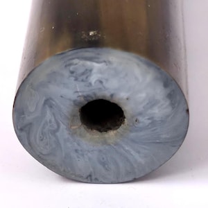 Puede incluir: Un objeto cilíndrico con una superficie exterior lisa de color marrón oscuro. La cara circular presenta un patrón jaspeado de tonos grises y blancos arremolinados, con una abertura circular oscura y centrada que se extiende hacia el interior.