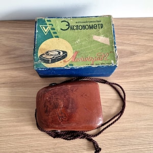 May include: Vintage light meter with a brown leather case and a box. The box is blue and green with Cyrillic text and a diagram of the meter. The leather case has a brown cord attached.