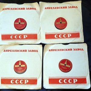 Puede incluir: Cuatro etiquetas de discos de vinilo blancas con letras rojas y doradas. Las etiquetas dicen "АПРЕЛЕВСКИЙ ЗАВОД" y "СССР". Cada etiqueta presenta un círculo rojo con una representación de un edificio.