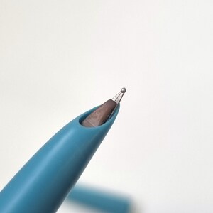 Puede incluir: Primer plano de un bol&iacute;grafo turquesa con punta de bola plateada. El cuerpo del bol&iacute;grafo es de color turquesa liso y la punta es plateada met&aacute;lica. El bol&iacute;grafo est&aacute; sobre un fondo blanco liso.