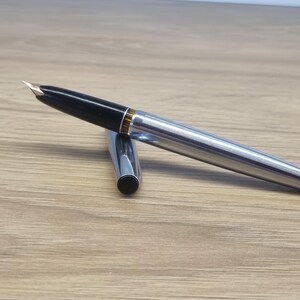 Puede incluir: Una pluma estilogr&aacute;fica plateada con capuch&oacute;n negro y plum&iacute;n dorado. El cuerpo del bol&iacute;grafo tiene un dise&ntilde;o acanalado. La tapa est&aacute; separada y colocada junto al bol&iacute;grafo. El bol&iacute;grafo est&aacute; sobre una superficie de madera.