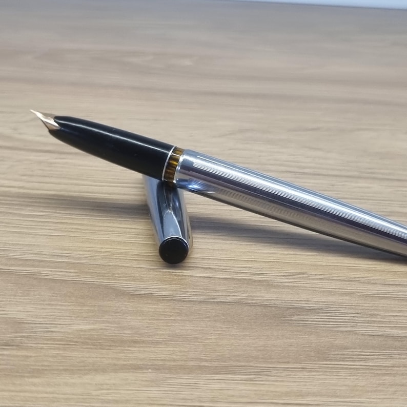 Puede incluir: Una pluma estilogr&aacute;fica plateada con capuch&oacute;n negro y plum&iacute;n dorado. El cuerpo del bol&iacute;grafo tiene un dise&ntilde;o acanalado. La tapa est&aacute; separada y colocada junto al bol&iacute;grafo. El bol&iacute;grafo est&aacute; sobre una superficie de madera.