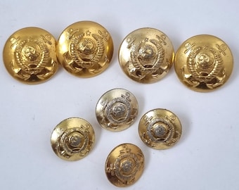 May include: A collection of seven circular gold-toned metal buttons featuring a raised emblem of a globe surrounded by a wreath and topped with an anchor. The buttons vary in size, with four larger buttons arranged at the top and three smaller ones below.