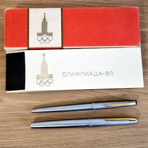 Może przedstawiać: Dwa srebrne długopisy ze złotymi akcentami i ich oryginalne pudełka. Pudełka są czerwono-białe ze złotym logo olimpijskim i napisem "ОЛИМПИАДА-80". Długopisy mają elegancki i nowoczesny design.