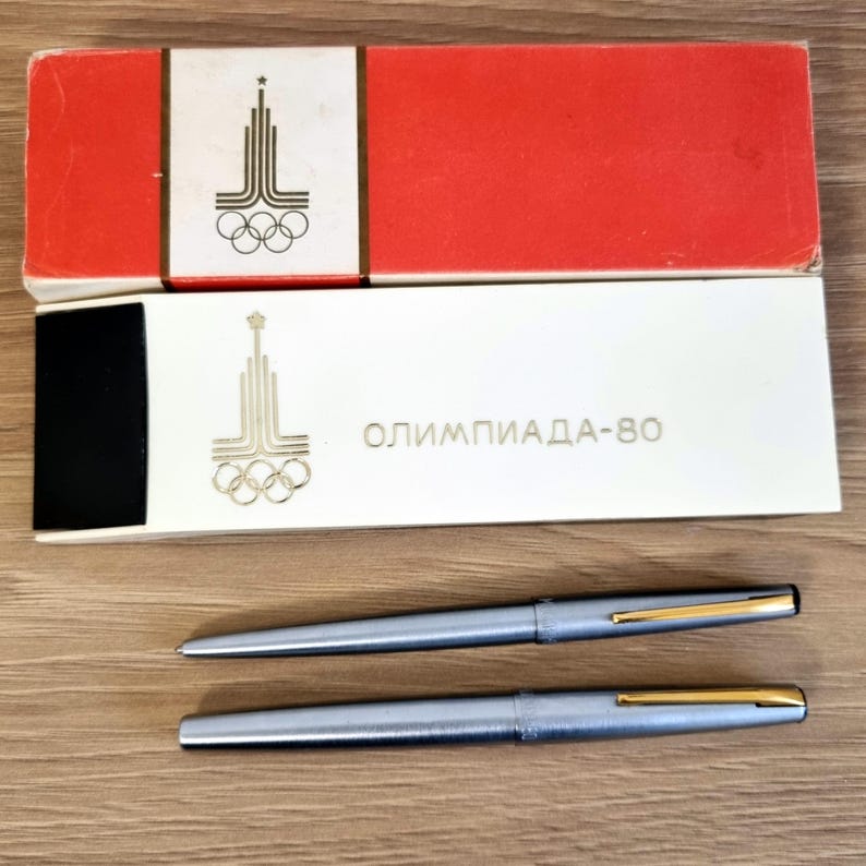 Może przedstawiać: Dwa srebrne długopisy ze złotymi akcentami i ich oryginalne pudełka. Pudełka są czerwono-białe ze złotym logo olimpijskim i napisem "ОЛИМПИАДА-80". Długopisy mają elegancki i nowoczesny design.
