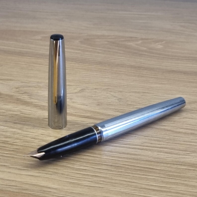 Puede incluir: Una pluma estilogr&aacute;fica plateada con agarre negro y plum&iacute;n dorado. La tapa est&aacute; quitada y colocada junto a la pluma. La pluma est&aacute; sobre una superficie de madera.