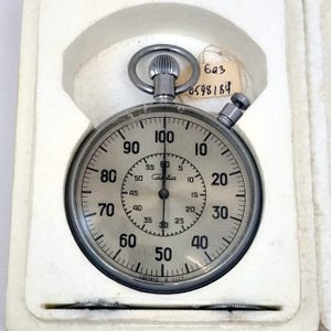 May include: A vintage silver stopwatch with a round face, featuring black numerals and markings against a light grey background. The stopwatch is in its original white case. The face displays numbers from 0 to 100, with smaller increments for seconds.