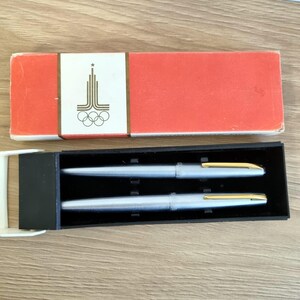 Może przedstawiać: Zestaw dw&oacute;ch srebrnych długopis&oacute;w ze złotymi akcentami, prezentowany w czarnym etui. Długopisy są pokazane obok czerwonego pudełka z białą etykietą z wystylizowanym emblematem i pierścieniami olimpijskimi.