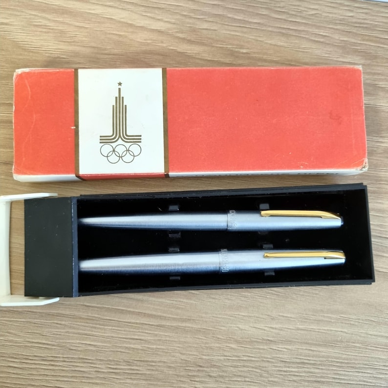 Może przedstawiać: Zestaw dw&oacute;ch srebrnych długopis&oacute;w ze złotymi akcentami, prezentowany w czarnym etui. Długopisy są pokazane obok czerwonego pudełka z białą etykietą z wystylizowanym emblematem i pierścieniami olimpijskimi.